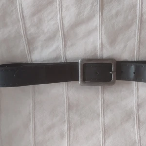 Svart/grått skärp/bälte med metallspänne - Grått/svart bälte i vad jag tror är nån typ av fakeskinn med ett rejält fyrkantigt metallspänne. Bältet har flera hål för justering och en stilren, enkel design som passar till jeans eller byxor. Perfekt för dig som gillar en klassisk look med lite edge. Ca 100 cm långt. Har bara använts ett fåtal ggr.