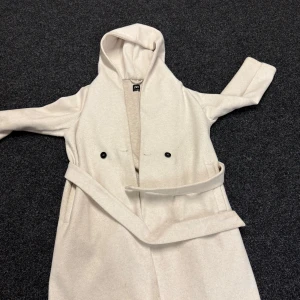 Beige kappa med huva från Zara - Snygg beige kappa från Zara med huva och bälte i midjan. Jackan har långa ärmar, två svarta knappar framtill och en stilren, rak passform. Perfekt för dig som gillar minimalistisk stil och vill ha något som håller dig varm under kyliga dagar.