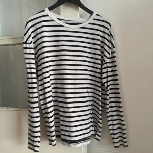 Randig långärmad tröja  - Snygg långärmad tröja i vit och svart med klassiska horisontella ränder. Tröjan är gjord i mjuk bomull och har en rund halsringning samt små slitsar i nederkanten. Perfekt för dig som gillar enkel och stilren design.