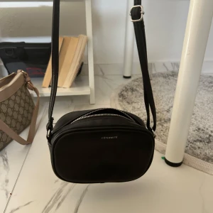 Svart axelväska från L'Éternité - Minimalistisk svart axelväska från L'Éternité i slät skinnimitation. Väskan har oval form, justerbar axelrem och silverfärgad dragkedja. Perfekt storlek för mobil, plånbok och nycklar. Stilren design med diskret logga framtill.