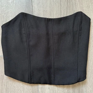 Svart korsett-topp från NA-KD - Snygg svart korsett-topp från NA-KD i storlek 34. Toppen har en stilren design med markerade sömmar framtill och smockad rygg för extra komfort. Axelbandslös och croppad modell som ger en trendig siluett. Perfekt att styla med höga jeans eller kjol.