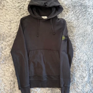 Svart hoodie från Stone Island - Ytterst lite solblekt! Svart hoodie från Stone Island med klassisk patch på vänster ärm. Tröjan har huva med snörning och en stor magficka framtill. Tillverkad i mjuk bomull, perfekt för chill dagar. Snygg och enkel design som funkar till det mesta.