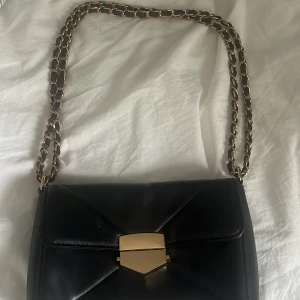 Svart axelväska med guldspänne och kedja - Snygg svart axelväska med mjukt skinnmaterial och guldfärgad kedja som axelrem. Väskan har ett geometriskt lock med ett coolt guldspänne framtill och en praktisk dragkedja på baksidan. Perfekt storlek för mobil, plånbok och småsaker. Bra skick
