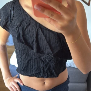 Svart oneshoulder topp från MNG - Snygg svart croppad topp från MNG med oneshoulder-design och tunna glittriga ränder. Materialet är lätt och har en ribbad känsla, perfekt för dig som vill sticka ut med en cool asymmetrisk look. Passformen är normal och toppen är ärmlös på ena sidan.