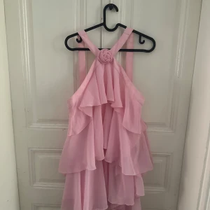 Ljusrosa volangklänning med rosett - Supersöt ljusrosa klänning med flera lager volanger och en dekorativ rosett framtill. Klänningen har axelband som korsas i ryggen och är tillverkad i ett lätt, flödande material som ger en romantisk vibe. Perfekt för sommarens alla tillfällen!