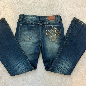 Blå bootcut jeans med guldnitar - Snygga blå jeans med bootcut passform och coola slitningar framtill. Baksidan har en bakficka dekorerad med guldiga nitar i ett mönster som ger extra edge. Klassisk femficksmodell och brunt läderpatch i midjan. Perfekta för dig som gillar detaljer och stil. Midjemåttet är 37cm och innebenslängden 82 cm.