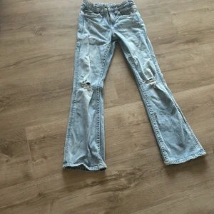 Bootcut ljusblå jeans med slitningar - Säljer ett par ljusblå bootcut jeans med snygga slitningar vid knäna och klassiska fickor bak med knappdetaljer. Jeansen har en låg midja och är tillverkade i mjukt denimtyg som ger en chill vibe. Perfekt för dig som gillar retro och avslappnad stil. Priset kan diskuteras. OPS 59 kr för frakten.