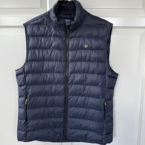 Mörkblå dunväst från Morris - Snygg mörkblå dunväst från Morris med quiltad design och ståkrage. Västen har dragkedja framtill, två sidofickor med gulddetaljer och en liten Morris-logga på bröstet. I princip aldrig använd så i perfekt skick. 