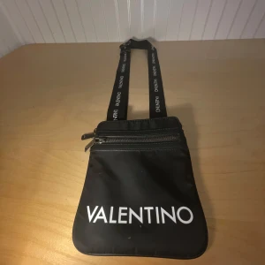 Svart axelväska från Valentino - Cool svart axelväska från Valentino med justerbar rem där loggan är tryckt i vitt. Väskan har två dragkedjefack och är tillverkad i slitstark polyester. Perfekt storlek för mobil, plånbok och småsaker. Snygg och enkel design med vita detaljer. Och sist men inte minst 100% äkta