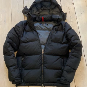 Svart dunjacka från Polo Ralph Lauren - Säljer en svart dunjacka från Polo Ralph Lauren. Jackan har en praktisk huva och dragkedja framtill. Perfekt för kyliga dagar med sin pufferdesign som håller värmen. Klassisk, stilren och trendig look. Pris kan diskuteras!