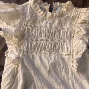Vit blus från Holly & Whyte - Säljer en söt vit blus från Holly & Whyte med vackra spetsdetaljer och volanger på ärmarna. Blusen har en rund hals och en guld knapp i ryggen. Köpt för ca 400kr💗