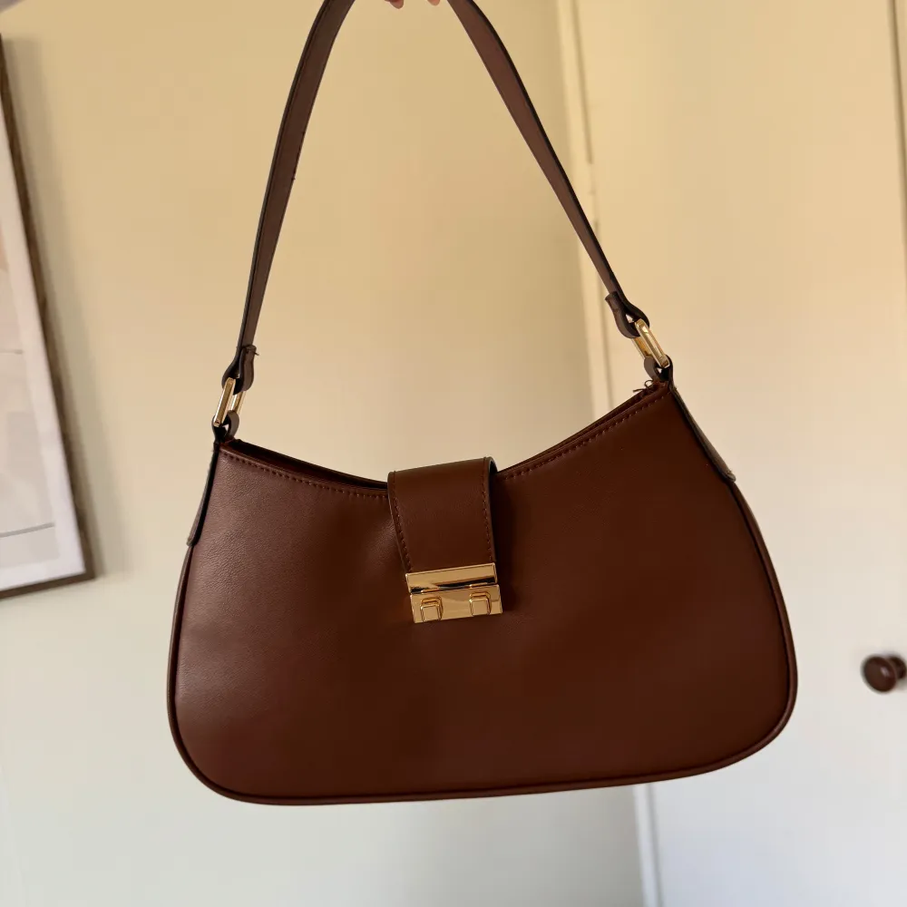 Snygg brun axelväska i skinn med en elegant gulddetalj på framsidan. Väskan har en stilren design med en enkel rem och en praktisk storlek för vardagsbruk. Perfekt för att ge din outfit en touch av klass.. Laukut & Käsilaukut.