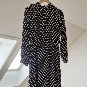Svart prickig klänning - Snygg svart klänning med vita prickar och långa ärmar. Perfekt för en stilren look med knappar framtill och en bekväm passform.