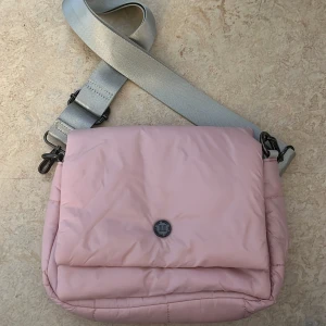 Rosa axelväska med bred rem - Snygg rosa axelväska med en bred, justerbar grå rem. Väskan är av hög kvalitet och fungerar även perfekt som makeup-väska. Stilren design och en fin färg som passar till allt!