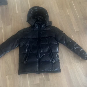 Svart pufferjacka med luva  - Säljer en svart pufferjacka med huva. Jackan har en glansig yta och dragkedja framtill samt fickor med dragkedjor. Perfekt för kyliga dagar när du vill hålla dig varm och stilren.