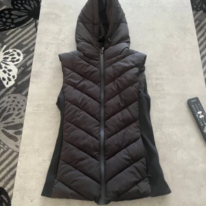 Svart dunväst från Zara - Snygg svart dunväst från Zara med huva och dragkedja framtill. Perfekt för lager-på-lager under kyliga dagar. Västen har en stilren design med quiltade detaljer som ger en modern look.