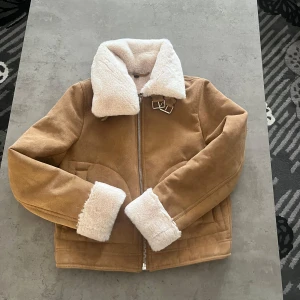 Beige jacka med fuskpäls från H&M Divided - Säljer en snygg beige jacka från H&M Divided med vit fuskpäls på krage och ärmslut. Jackan har en dragkedja framtill och dekorativa spännen vid kragen. Perfekt för kyliga dagar!