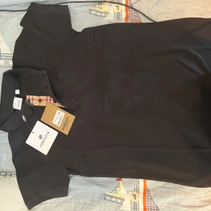 Svart pikétröja från Burberry - Säljer en stilren svart pikétröja från Burberry med klassisk krage och knappar. Tröjan har ett diskret broderat märke på bröstet och ikoniskt rutigt mönster på insidan av knappslån. Skriv gärna vid fler frågor/funderingar. 