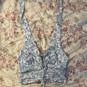 Blommig v-ringad topp - Säljer en söt blommig topp med v-ringning och knytning framtill. Toppen har ett blått blommönster på vit bakgrund och är croppad med smockdetaljer på baksidan. Perfekt för varma dagar!