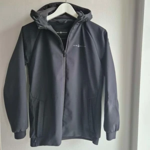 Svart jacka från Sail Racing - Snygg svart jacka från Sail Racing med dragkedja och huva. Perfekt för blåsiga dagar. Jackan har långa ärmar och en stilren design med logotyp på bröstet.