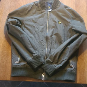 Grön bomberjacka från Lyle & Scott - Säljer en snygg grön bomberjacka från Lyle & Scott. Jackan har en dragkedja framtill och ribbade muddar vid ärmslut och nederkant. Den ikoniska loggan syns på bröstet. Perfekt för en stilren look!