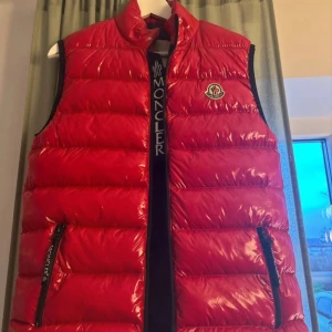 Röd dunväst från Moncler - Snygg röd dunväst från Moncler med dragkedja och logotyp på bröstet. Västen har en glansig finish och är perfekt för kyligare dagar. Den har en klassisk design med två fickor framtill.