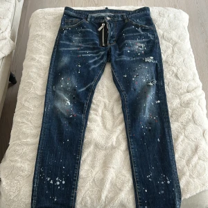 Mörkblå jeans med färgstänk från DSQUARED2 - Snygga mörkblå jeans från DSQUARED2 med unika färgstänk och slitna detaljer. Jeansen har en klassisk femficksdesign och en knappgylf. Perfekta för dig som vill ha en edgy look.