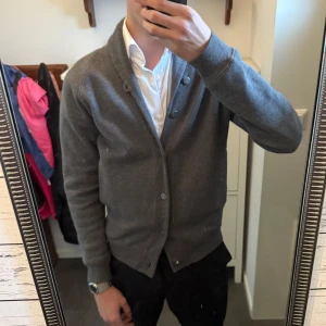 Grå kofta/Cardigan från J.Lindeberg - Säljer en stilren grå kofta från J.Lindeberg. Koftan har en mjuk insida och knappar framtill. Perfekt för kyligare dagar och passar bra över en skjorta. Bra skick. Storlek S