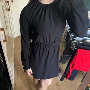 Klänning  - Säljer en stilren svart klänning med lång ärm och elastisk midja. Perfekt för en elegant look. Klänningen har en knappdetalj i nacken och är gjord i ett mjukt material som ger en bekväm passform.