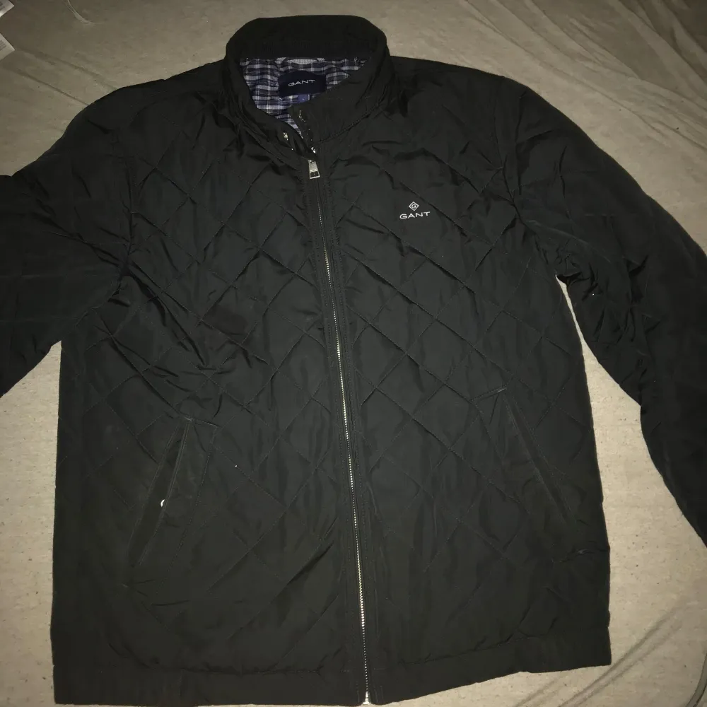 Säljer en stilren svart quiltad jacka från GANT. Jackan har en dragkedja framtill och ett rutigt foder i blått och brunt. Perfekt för kyligare dagar med sin klassiska design.(PRIS KAN DISKUTERAS). Takit.