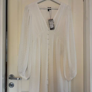 Vit klänning från Vero Moda - Säljer en fin vit klänning från Vero Moda med v-ringning och långa ärmar. Klänningen har knappar framtill och ett diskret prickigt mönster. Klänningen är helt ny och är endast provad, perfekt midsommar- eller studentklänning. Storlek 38/M