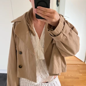 Trenchcoat  - Säljer en snygg trenchcoat från Gina Tricot. Den har en croppad design med stora knappar och långa ärmar. Superfin nu till våren. Det är en storlek L men är liten i storleken så passar mig som är en s/m🤗