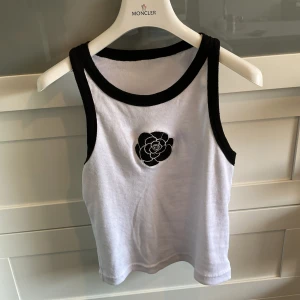 Vit ribbad linnetopp med svart blomdetalj - Säljer en snygg vit ribbad linnetopp från Moncler med svarta kanter och en cool svart blomdetalj på bröstet. Perfekt för en stilren look!