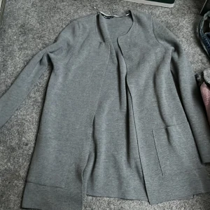 Grå långärmad cardigan från Gina Tricot - Säljer en stilren grå långärmad cardigan från Gina Tricot. Den har en öppen design utan knappar och är perfekt för lager-på-lager. Passar bra till både vardag och mer uppklädda tillfällen.