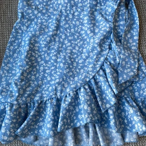 Blå blommig kjol - Söt blå kjol med vita blommor och volangdetaljer. Perfekt för sommarens alla tillfällen. Kjolen har en omlott-design som ger en fin passform. Jättebra skick, som ny. Storlek: XS men passar både större och mindre