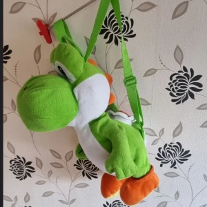 Yoshi ryggsäck, Nintendo, original, presenter, julklapp, mario, animations - Säljer en unik grön axelväska i form av en dinosaurie Yoshi. Väskan är gjord av polyester och har en praktisk dragkedja på baksidan. Perfekt för att bära småsaker och ge en lekfull touch till din stil.Yoshi ryggsäck med förvaring i Yoshis skal.  Original Nintendo 2012, "Animations" New York. Mått: ca 45cm lång och 25cm bred.