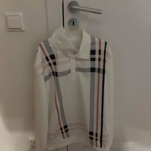 Vit långärmad tröja med rutigt mönster - Säljer en stilren vit långärmad tröja med ett klassiskt rutigt mönster i svart, beige och rosa. Tröjan har en hög krage och en avslappnad passform. Perfekt för en trendig och bekväm look.