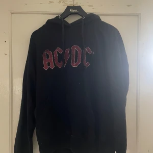 Svart AC/DC hoodie från House Brand - Säljer en svart hoodie med AC/DC-tryck från House Brand. Tröjan har en klassisk design med huva och dragsko, samt tryck på både fram- och baksidan med turnédatum. Perfekt för rockälskare! 🤘