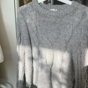 Grå stickad tröja från Gina Tricot - Säljer en mysig grå stickad tröja från Gina Tricot. Tröjan har ett klassiskt flätmönster och långa ärmar, perfekt för kyliga dagar. Den är mjuk och bekväm med ribbstickade muddar vid ärmslut och nederkant. Passar utmärkt för både höst och vinter.