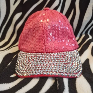 Glittrig rosa keps från Boland - Häftigt rosa keps från Boland med glittriga paljetter och en skärm täckt av silverfärgade nitar. Perfekt för att sticka ut på festen! Justerbar rem baktill för bästa passform. Använt typ en gång innan och på sista bilden kan man se att en av nitarna är lite sne men man märker knappt när den är på. 🩷 100% polyester.