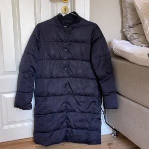 Svart dunjacka - Säljer en stilren svart dunjacka med dragkedja och knappar framtill. Jackan har en lång och rak passform. Perfekt för kyligare dagar.