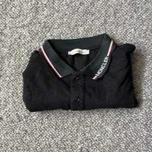 Moncler piké  - Snygg svart pikétröja med broderad text på kragen. Tröjan har en klassisk design med knappar framtill och ränder i rött och vitt på kragen. Perfekt för en stilren look. Inte helt säker på om den är äkta eftersom jag fick den av min bror.