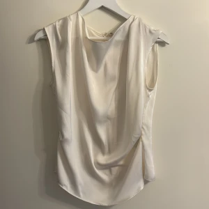 Vit ärmlös topp från H&M - Säljer en stilren vit ärmlös topp från H&M i ett mjukt och glansigt material. Toppen har en dragkedja i ryggen för enkel på- och avtagning. Perfekt för en elegant look.