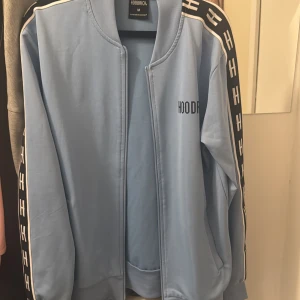 Ljusblå jacka från Hoodrich - Säljer en ljusblå kodta från Hoodrich med svarta och vita detaljer längs ärmarna. Kodtan har en dragkedja framtill och en stilren design med logotypen tryckt på bröstet. Perfekt för en avslappnad stil. Använd cirka 2 gr då den inte passade