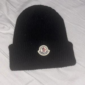 Svart ribbad mössa från Moncler - Snygg svart ribbad mössa från Moncler med broderad logga framtill. Perfekt för kyliga dagar och ger en stilren look. Tillverkad i ett mjukt och bekvämt material. Skick 10/10 aldrig använd