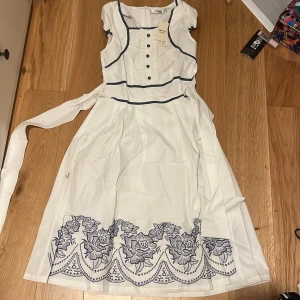 Vit klänning med blå broderier från Orfly XS - Charmig vit klänning med blå broderade blommor längs fållen och dekorativa knappar framtill. Klänningen har en knytning i midjan och är ärmlös med blå detaljer längs kanterna. Perfekt för en somrig look!