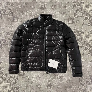 Svart pufferjacka från Moncler - Snygg svart pufferjacka från Moncler med glansig finish. Jackan har en dragkedja framtill och två praktiska fickor med dragkedja. Perfekt för kyliga dagar när du vill hålla stilen. 🖤