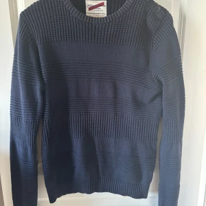 Mörkblå stickad tröja från Tailored Denim - Säljer en snygg mörkblå stickad tröja från Tailored Denim. Tröjan har ett randigt mönster med  knappar vid axeln. Perfekt för vår och sommarkvällar.