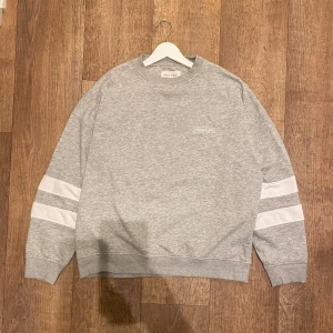Grå Sweatshirt från Pier One (Oversized) - Säljer en stilren grå sweatshirt från Pier One med vita ränder på ärmarna och ett subtilt tryck på bröstet. Tröjan har en oversized passform och långa ärmar, perfekt för en avslappnad look. Storlek Small. Snabb Leverans!