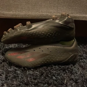 Fotbollsskor från Adidas - Snygga fotbollsskor från Adidas i en mörk färg med röda detaljer. Skorna har en strömlinjeformad design med dobbar för bra grepp på planen. Perfekta för fotbollsspelare som vill ha stil och funktion.